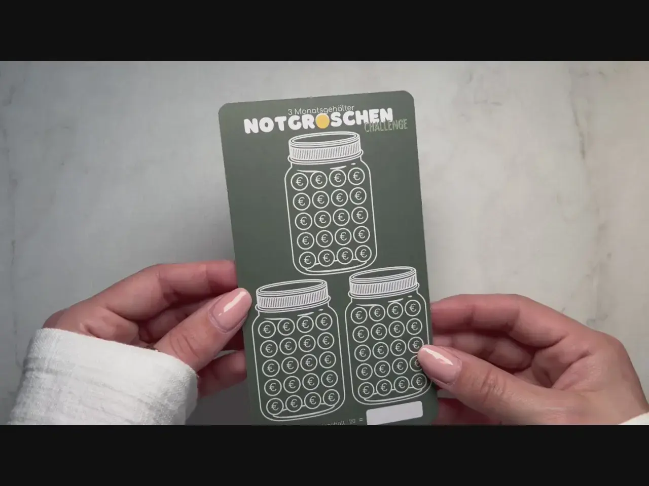 Hände halten eine "Notgroschen Challenge"-Karte mit drei Einmachgläsern, gefüllt mit Euro-Symbolen. Perfekt, um die eigene spar challenge selber machen zu starten!
