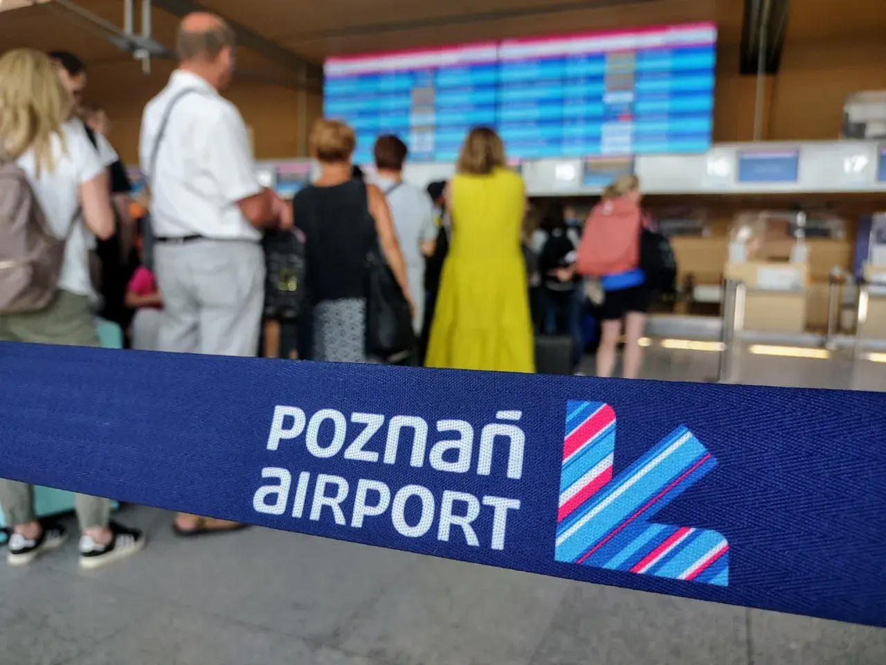Niebieska taśma z napisem "POZNAŃ AIRPORT" i logo lotniska na tle ludzi i tablicy odlotów.