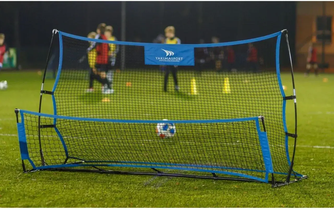 Piłkarski rebounder Yakimasport na zielonej murawie, z piłką w siatce. W tle trening młodych piłkarzy.