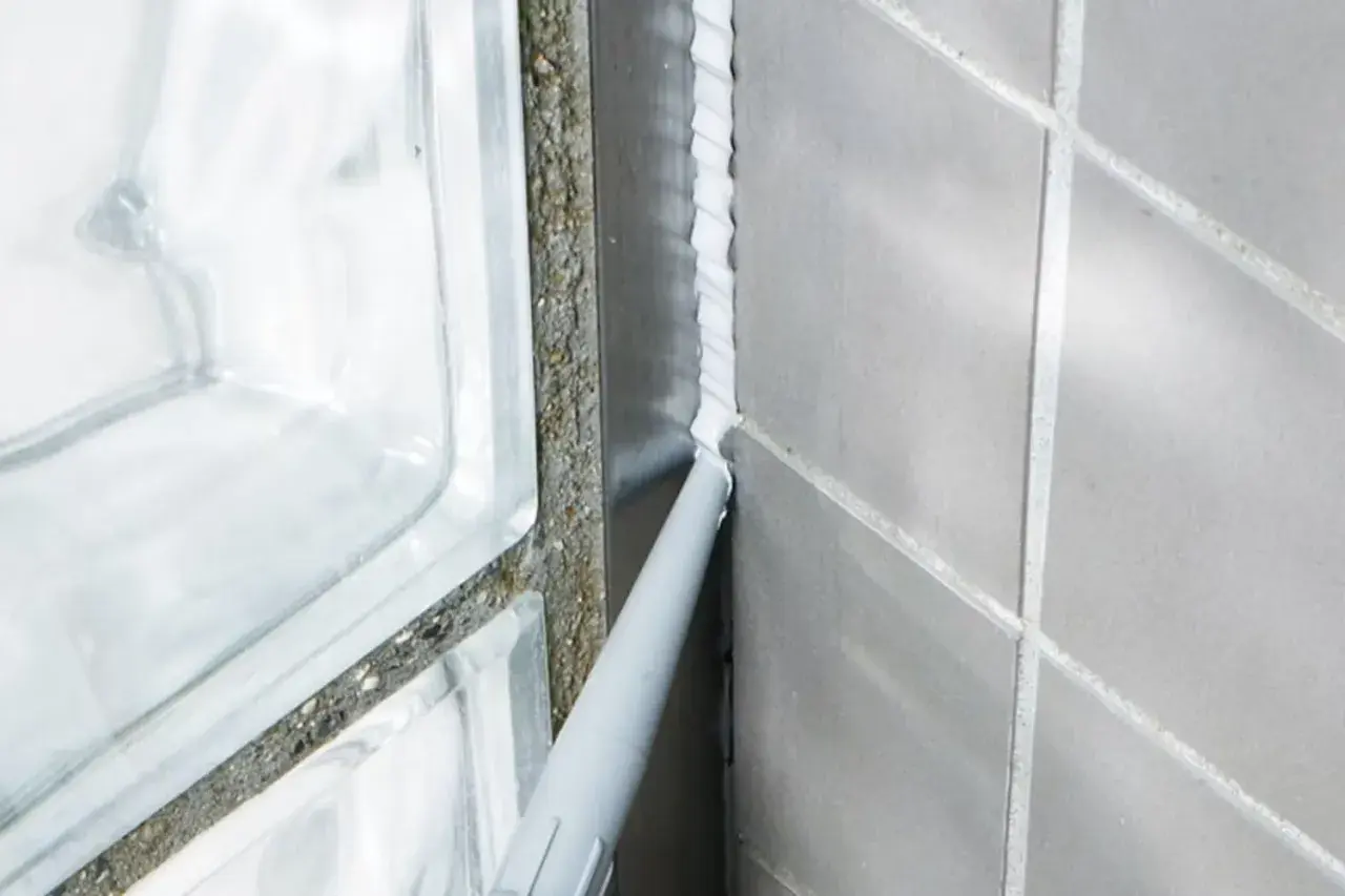Applicator sprutar vit silikonfog längs en fog i badrummet, intill en duschvägg av glasblock.