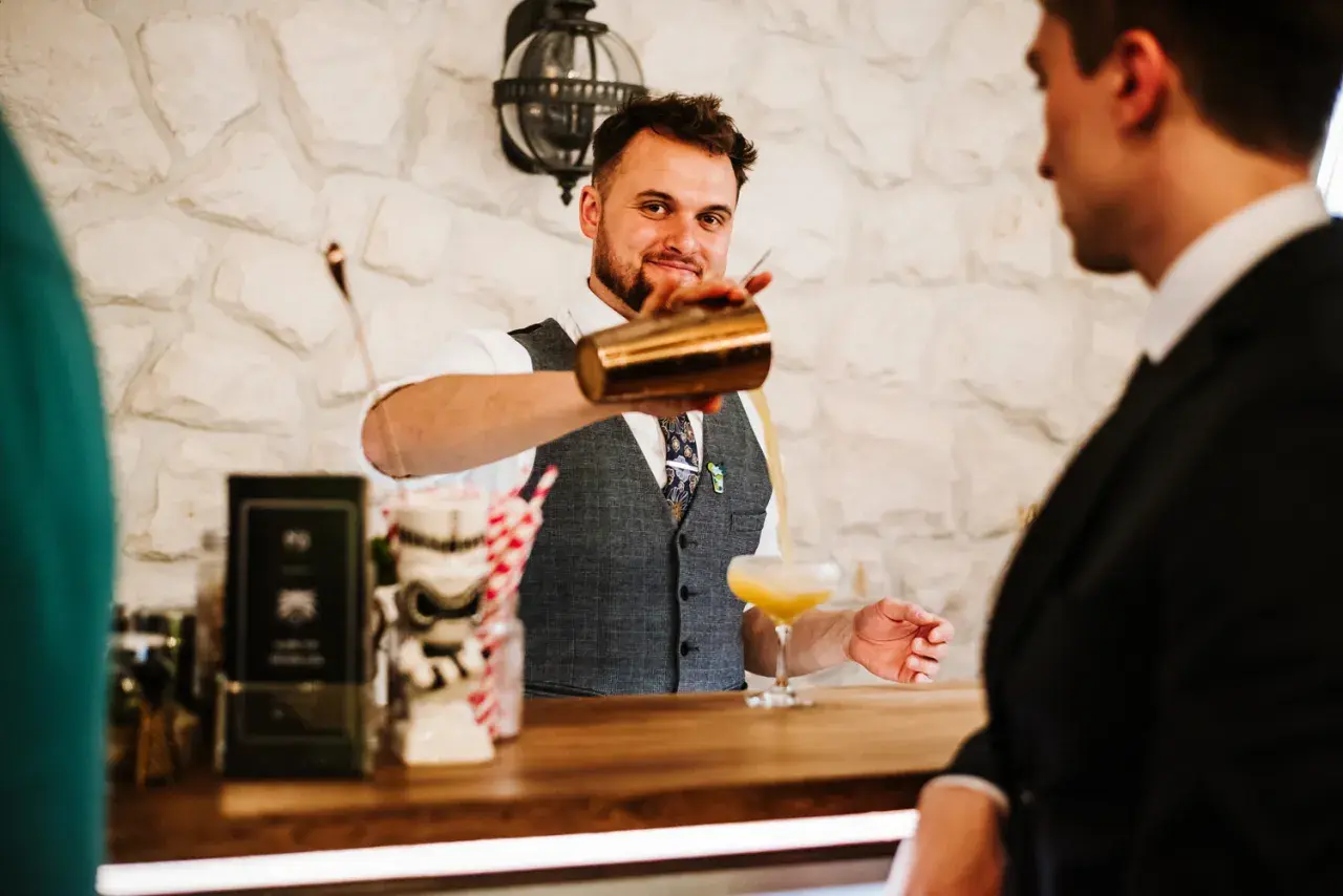 Uśmiechnięty barman w kamizelce nalewa drinka do kieliszka.