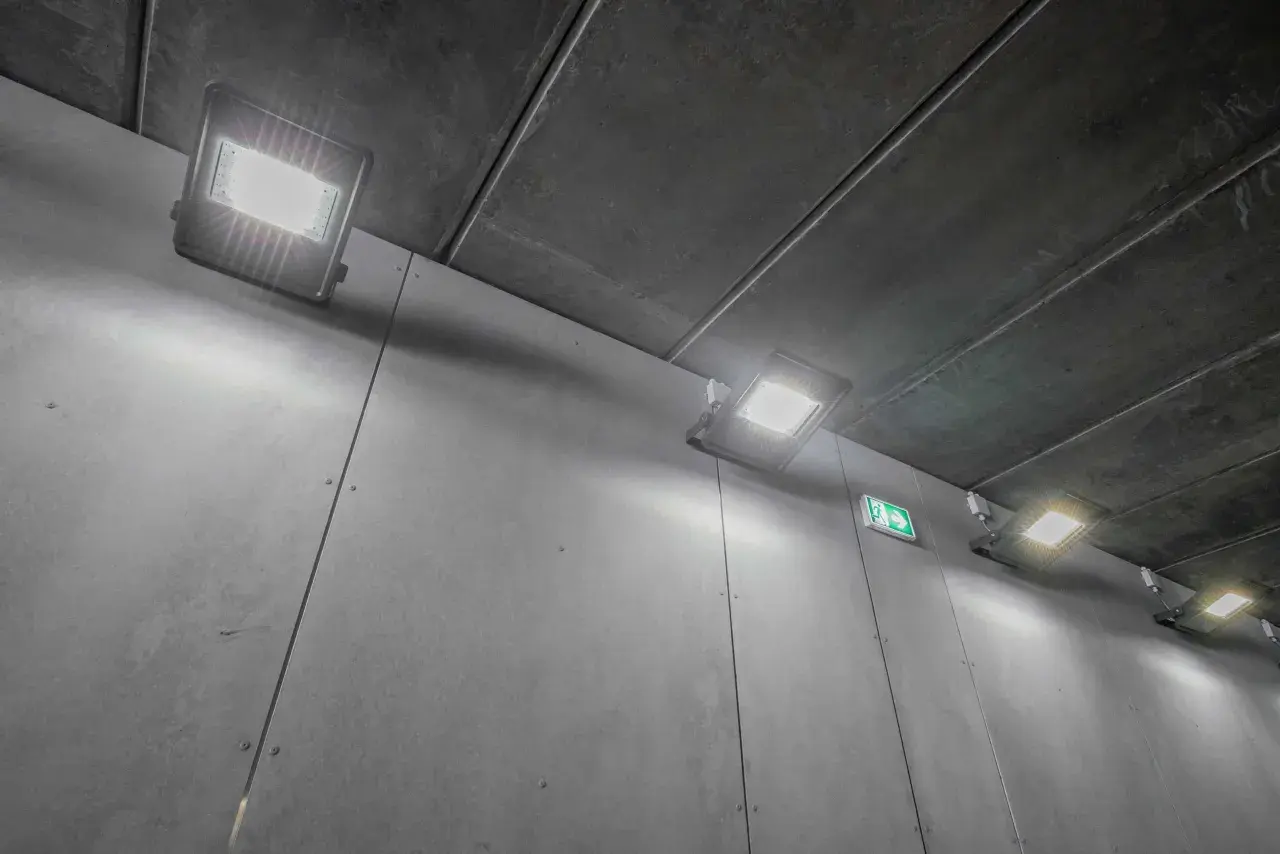 Jasne, oto opis alternatywny:

Oświetlenie długiego korytarza z betonowym sufitem i szarymi ścianami. Kilka nowoczesnych lamp LED rzuca jasne światło, a zielony znak ewakuacyjny wskazuje drogę.