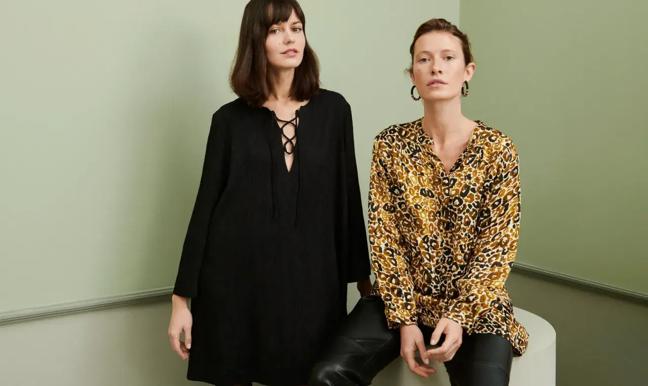 Två kvinnor poserar i en studio. Den ena bär en svart klänning med snörning, den andra en blus med leopardmönster. Båda utstrålar en elegant fransk stil.