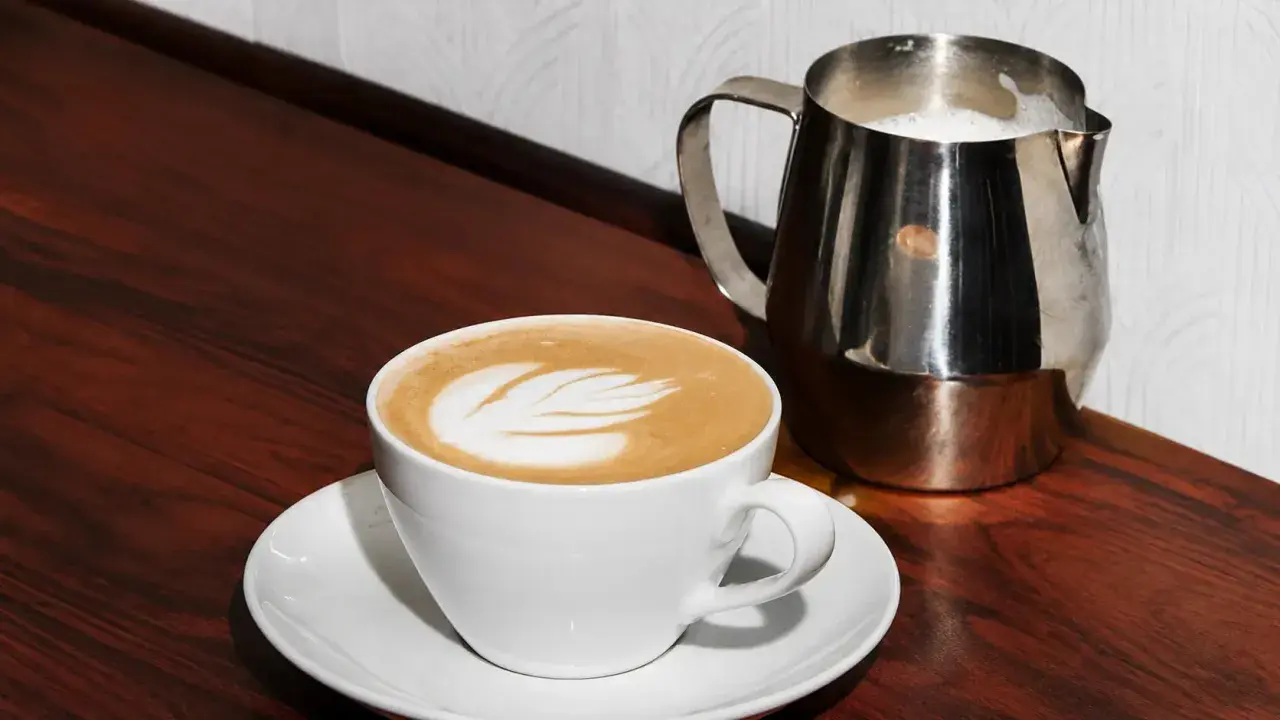 Biała filiżanka z latte art, obok metalowy dzbanek z mlekiem. Idealny raf coffee na drewnianym blacie.