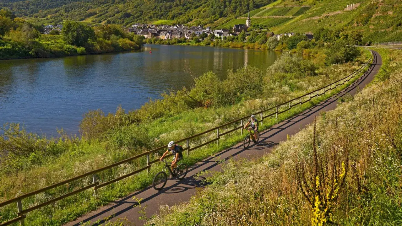 Zwei Radfahrer genießen die idyllischen radwanderwege deutschland entlang eines Flusses mit Weinbergen und einem Dorf im Hintergrund.