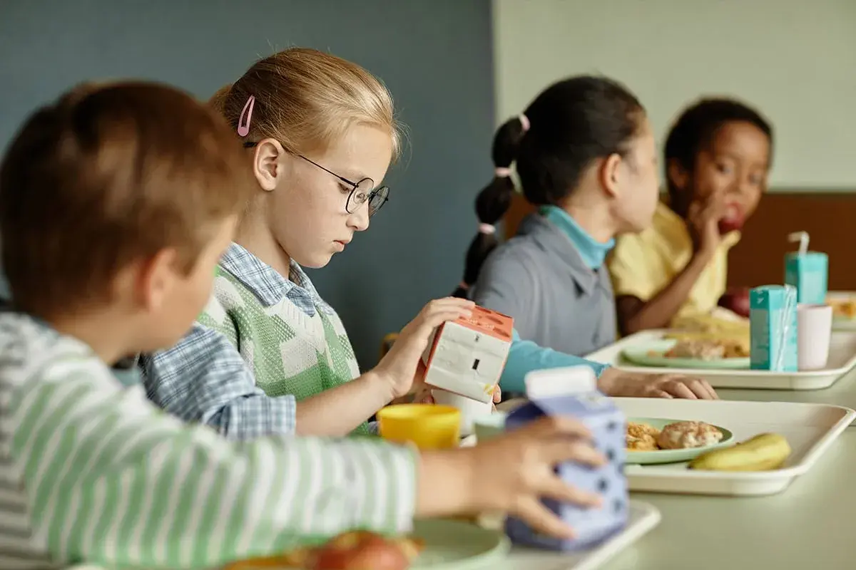 Kinder essen in der Mensa. Ein Mädchen mit Brille hält eine Milchpackung. Ein Teil des Tagesablaufs eines Schulbegleiters.