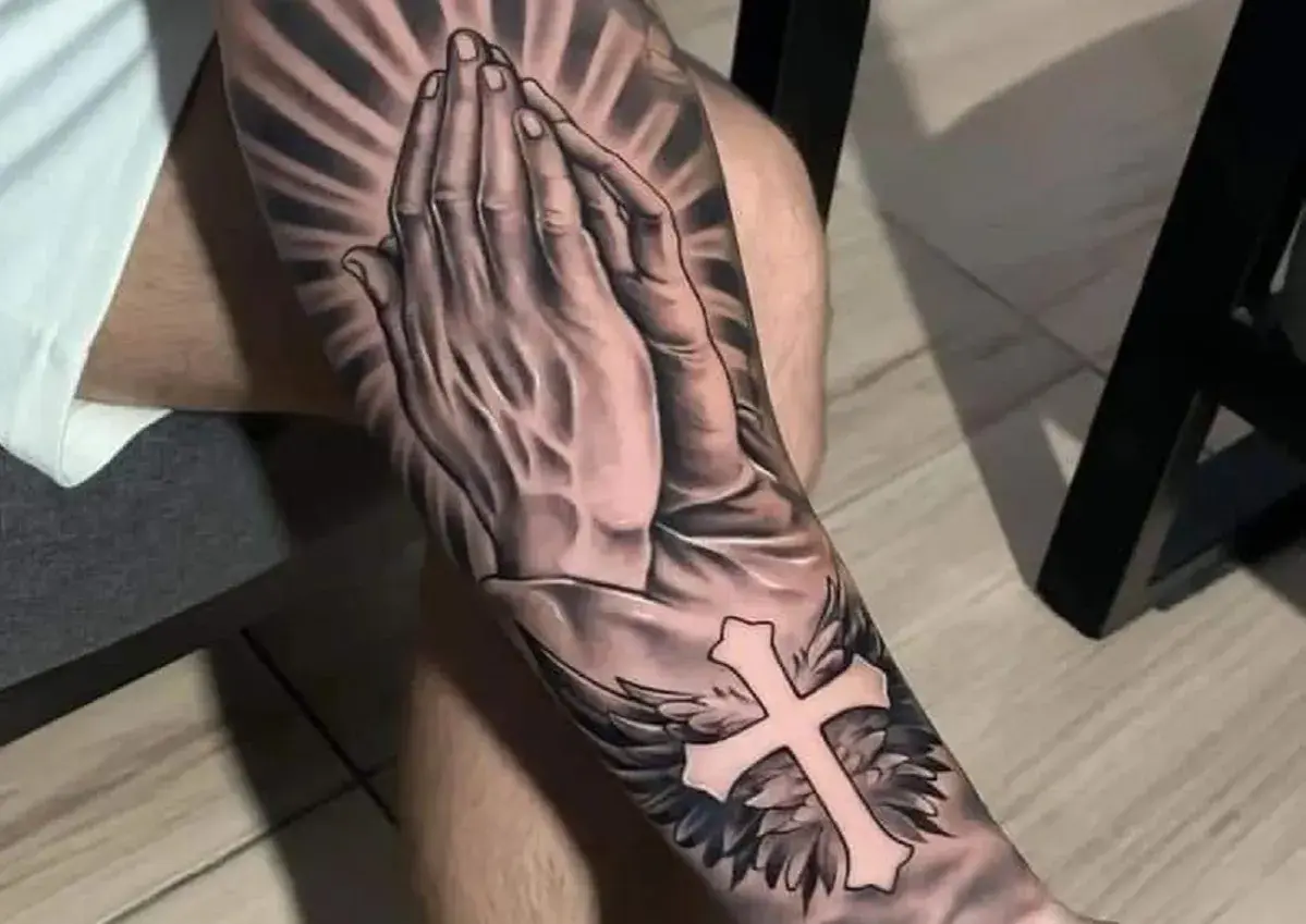 Tatuaż religijny mały: splecione dłonie w modlitwie, krzyż ze skrzydłami i promienie światła.
