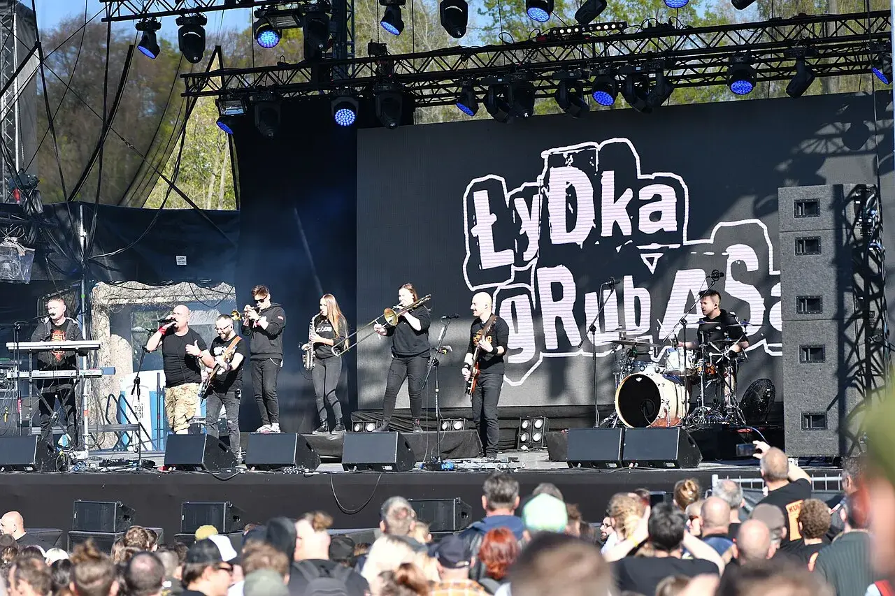 Zespół Łydka Grubasa na scenie, grający koncert. W tle wielki ekran z logo zespołu.