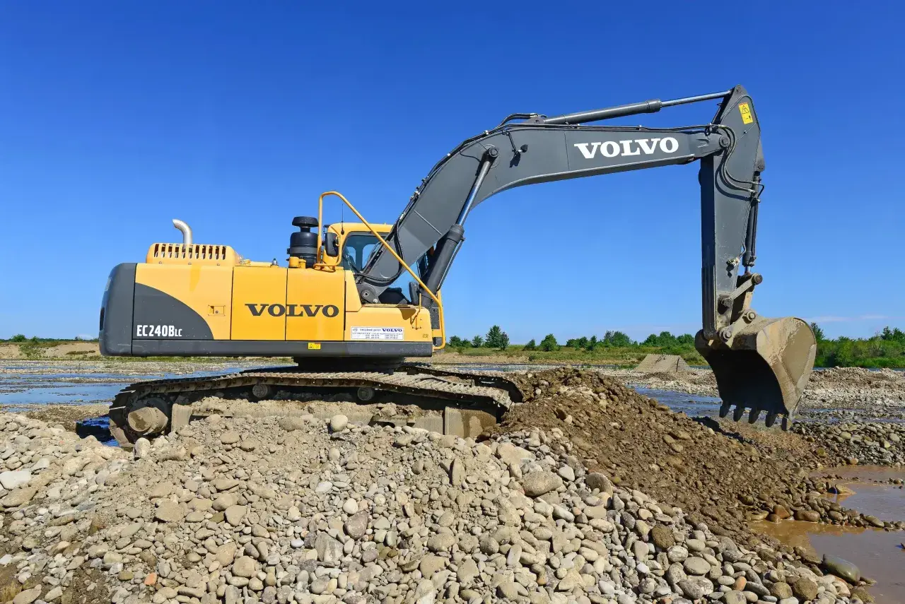 Żółta koparka Volvo EC240BLc pracuje na kamienistym brzegu rzeki. Zastanawiasz się, ile bierze koparka za godzinę?