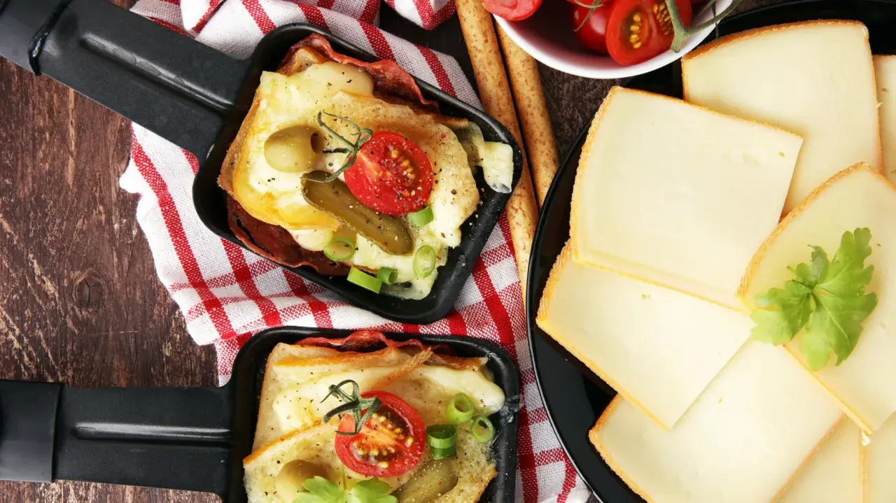 Deux poêlons de raclette fondue, garnie de tomates, cornichons et olives, accompagnés de fines tranches de fromage. Une variante gourmande !