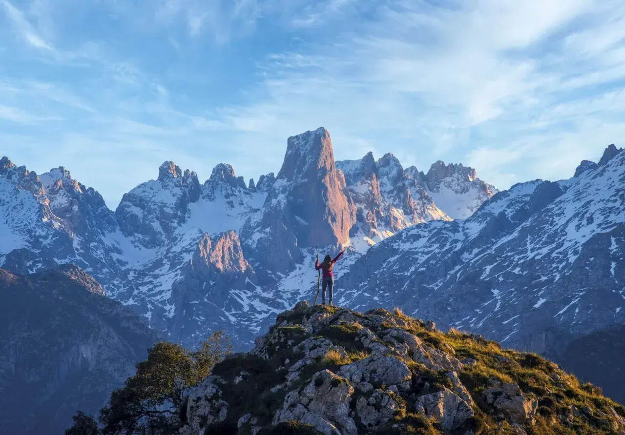 Persona en la cima de una montaña rocosa, con los brazos extendidos, contemplando los impresionantes picos nevados de los Picos de Europa. Un paisaje espectacular para descubrir qué ver.