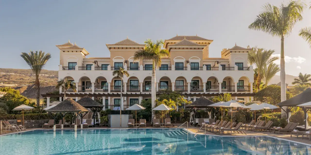 Piscina y edificio del Gran Meliá Palacio de Isora, con palmeras y tumbonas bajo el sol.
