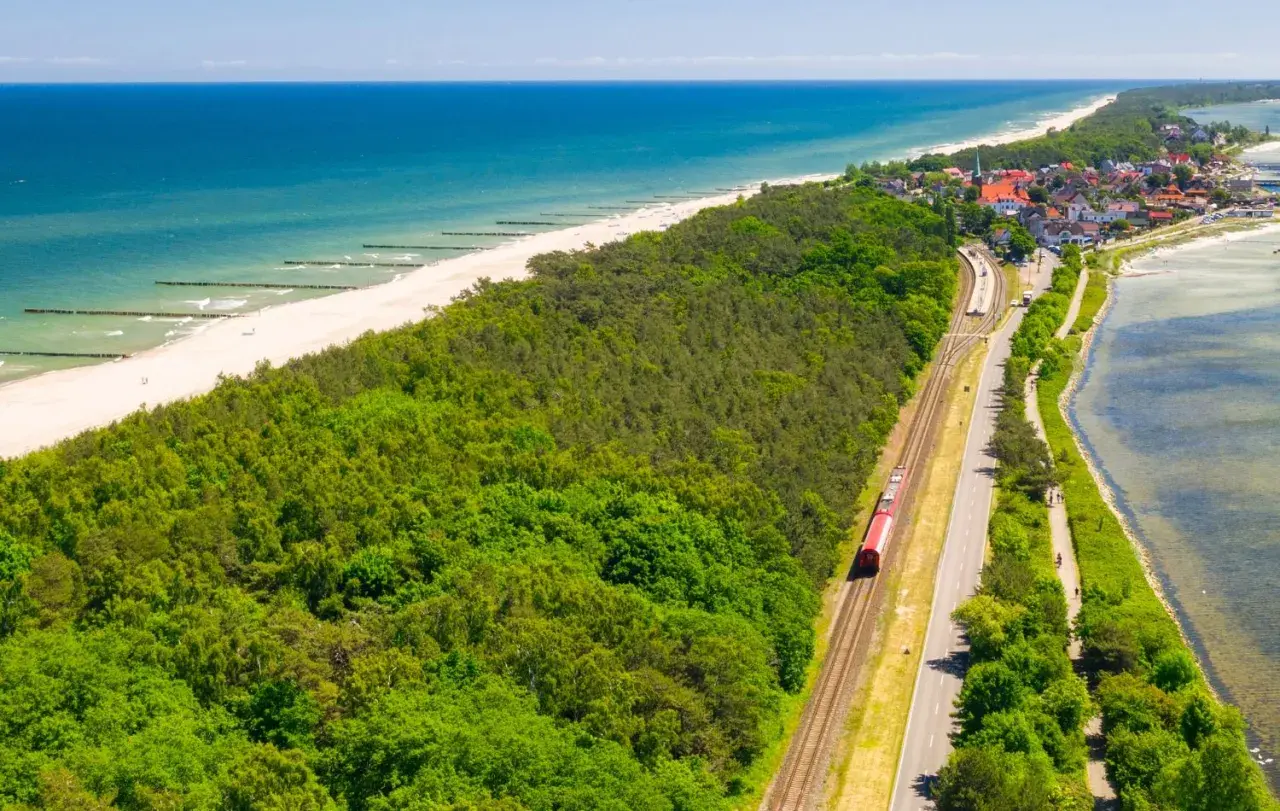 Czerwony pociąg jedzie wzdłuż wybrzeża. W tle widać plażę, morze i nadmorską miejscowość. Idealna podróż pociągiem nad morze.