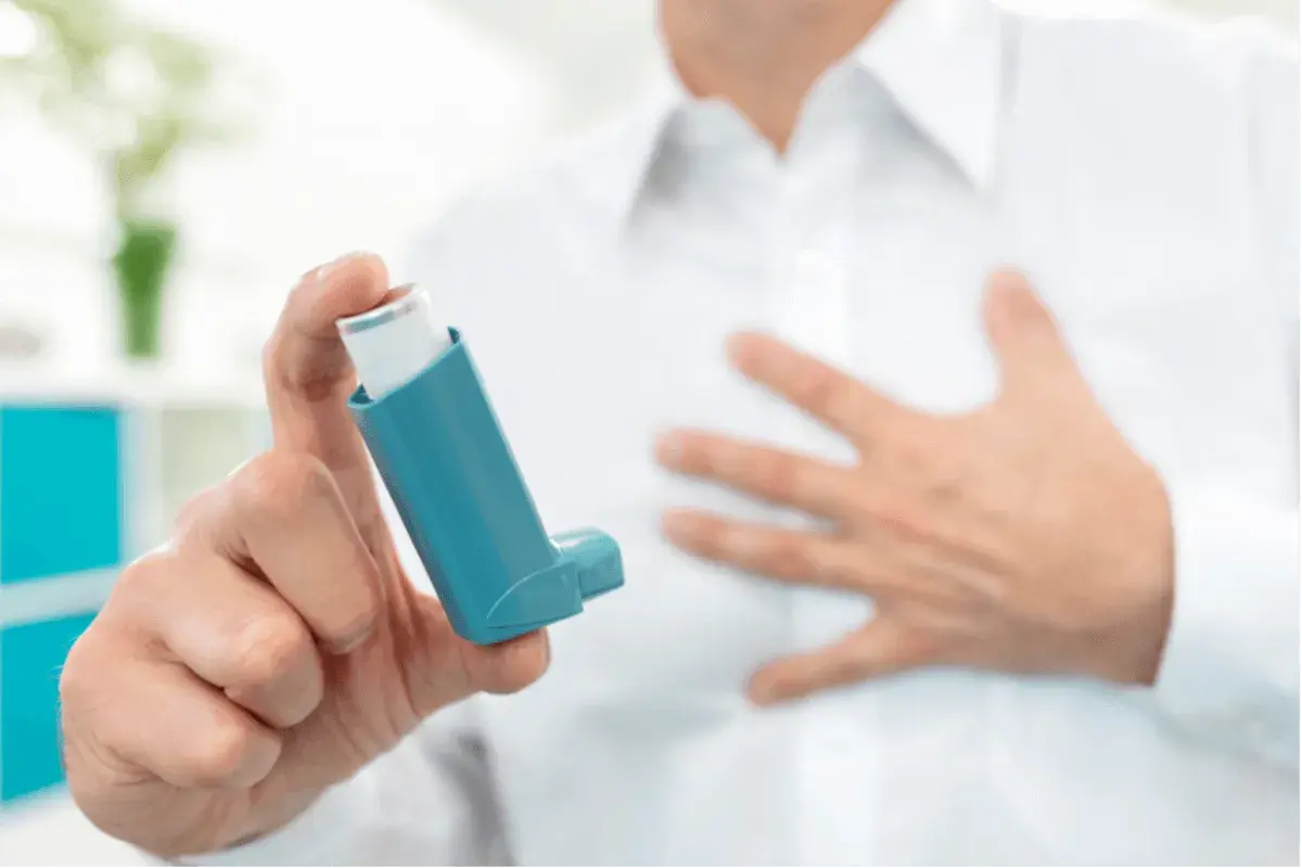 Mann hält Inhalator mit ipratropiumbromid. Seine andere Hand liegt auf der Brust, was auf Atemnot hindeutet.