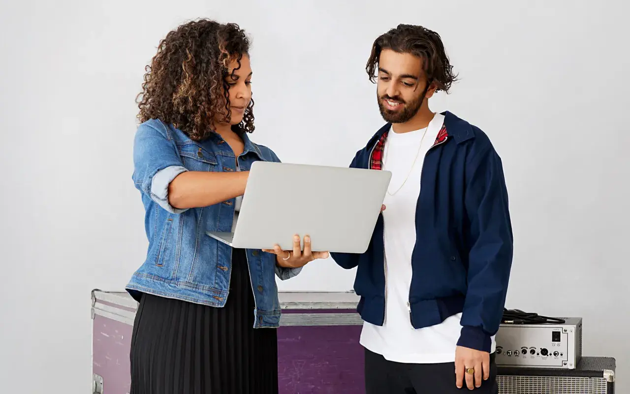 A woman and man review a laptop, discussing music artist management. They stand near stage equipment.