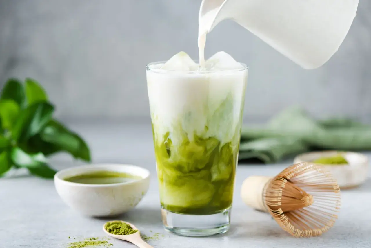 Eisiger Matcha Latte wird mit Milch übergossen. Die Zutaten für den Matcha Latte (kcal) sind auf dem Tisch verteilt.