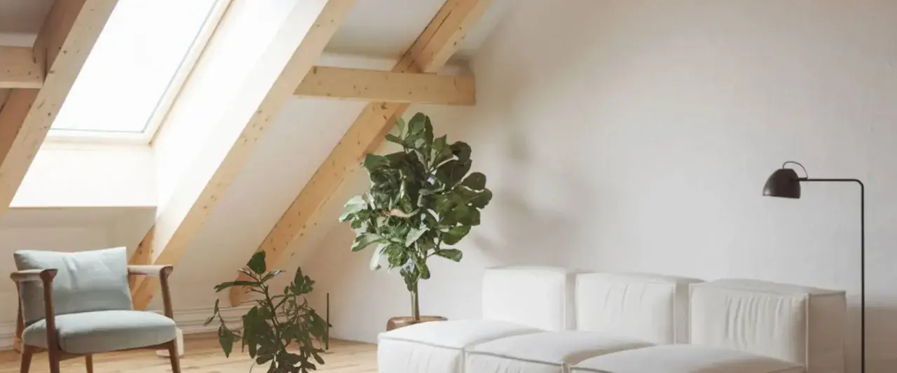 Un salon mansardé lumineux avec une fenêtre de toit, un fauteuil, des plantes et un canapé blanc. Idéal pour apprendre comment poser une fenêtre de toit.