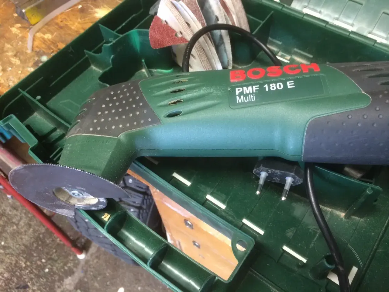 Bosch PMF 180 E multiverktyg med sågblad redo att kapa plasttak. Tillbehör och laddare ligger i lådan.