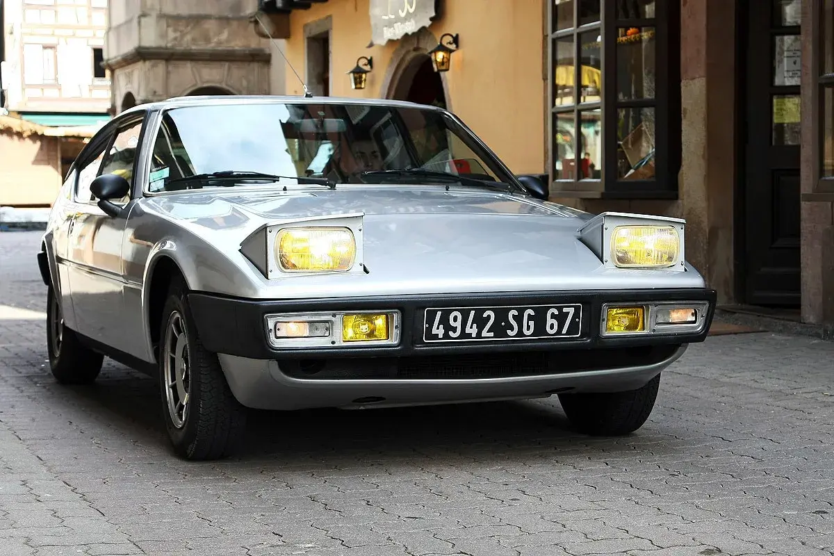Silberner Simca Bagheera mit ausklappbaren Scheinwerfern fährt durch eine Gasse mit Kopfsteinpflaster.