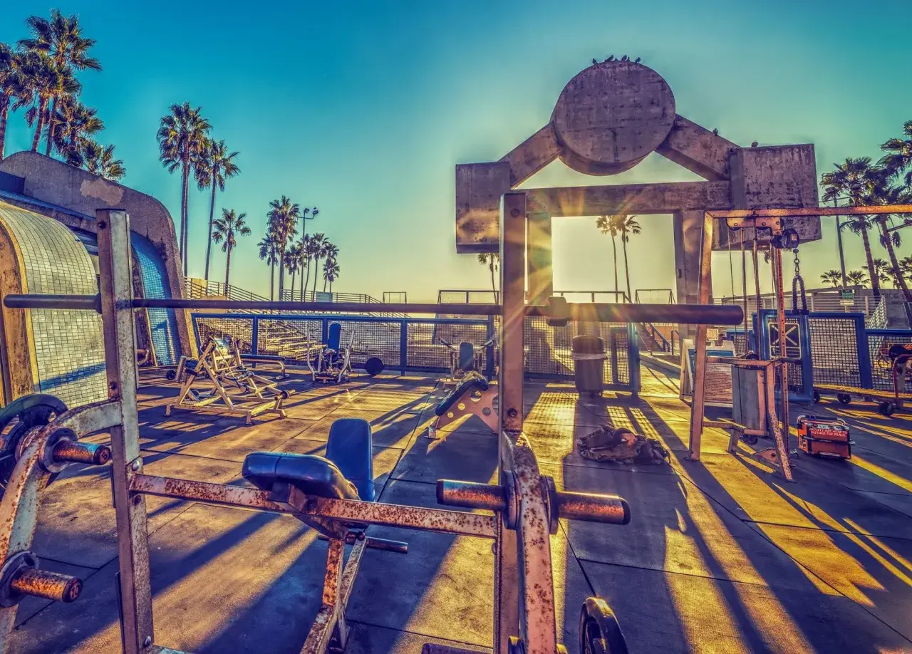 Słoneczny poranek na Venice Beach. Siłownia pod gołym niebem z palmami w tle. Długie cienie sprzętu do ćwiczeń na betonie.