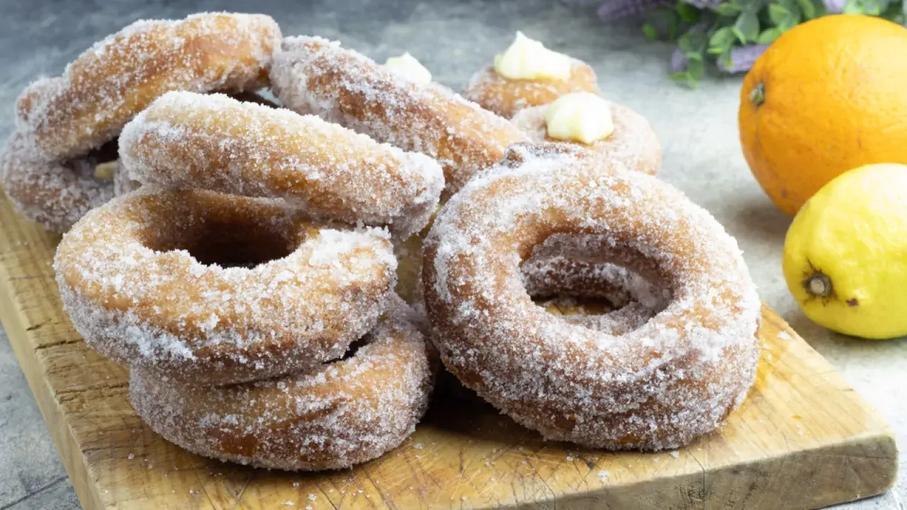 Un mucchio di ciambelle di carnevale zuccherate, alcune con crema, su un tagliere di legno, con arancia e limone sullo sfondo.