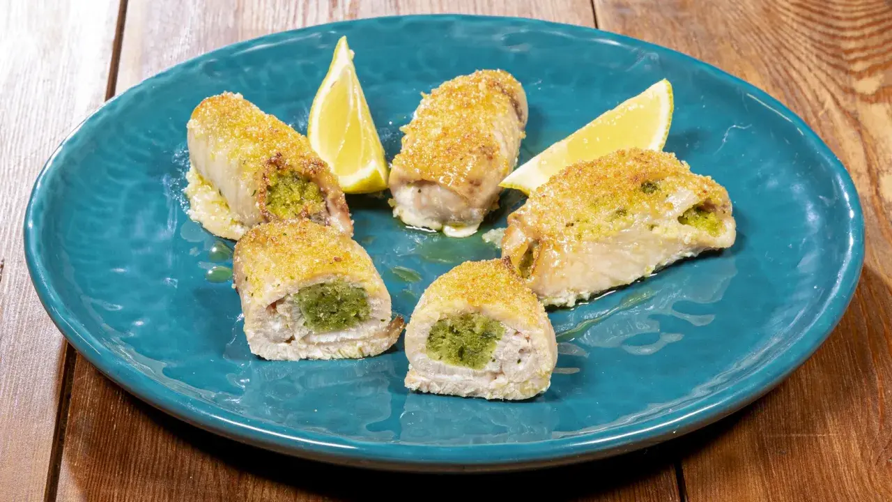 Involtini di pesce spada ripieni di erbe aromatiche, dorati e serviti su un piatto blu con spicchi di limone.