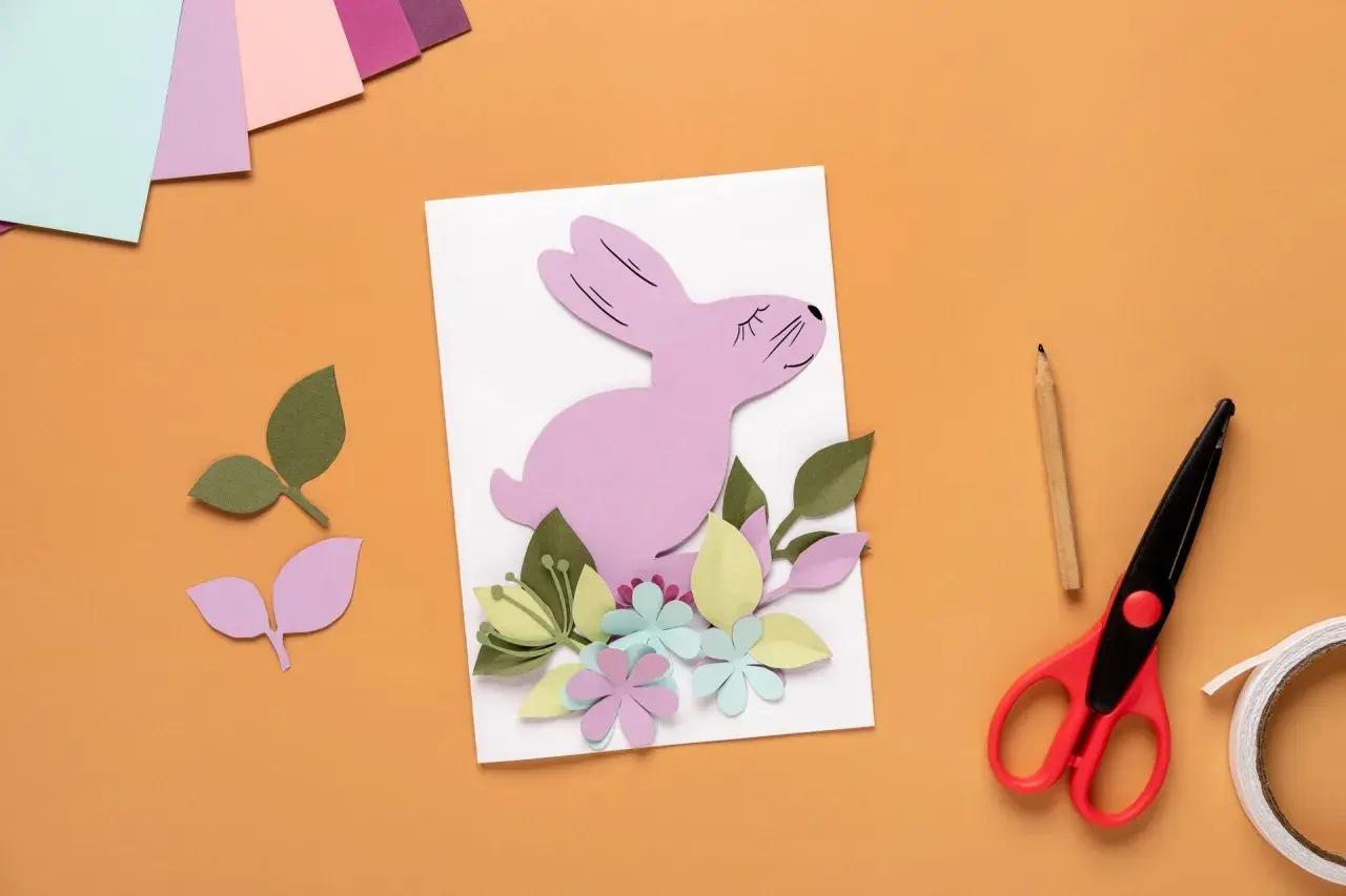 Uroczy różowy króliczek z papieru na kartce wielkanocnej DIY, otoczony kolorowymi kwiatami i liśćmi. Obok nożyczki, ołówek i taśma.