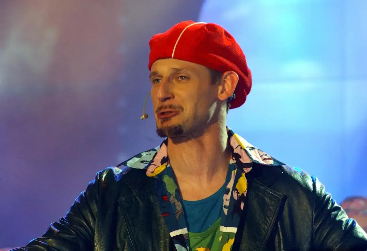 Michał Wójcik w czerwonej beretce i kolorowej koszuli, z mikrofonem przy ustach, na tle niebiesko-fioletowego światła.