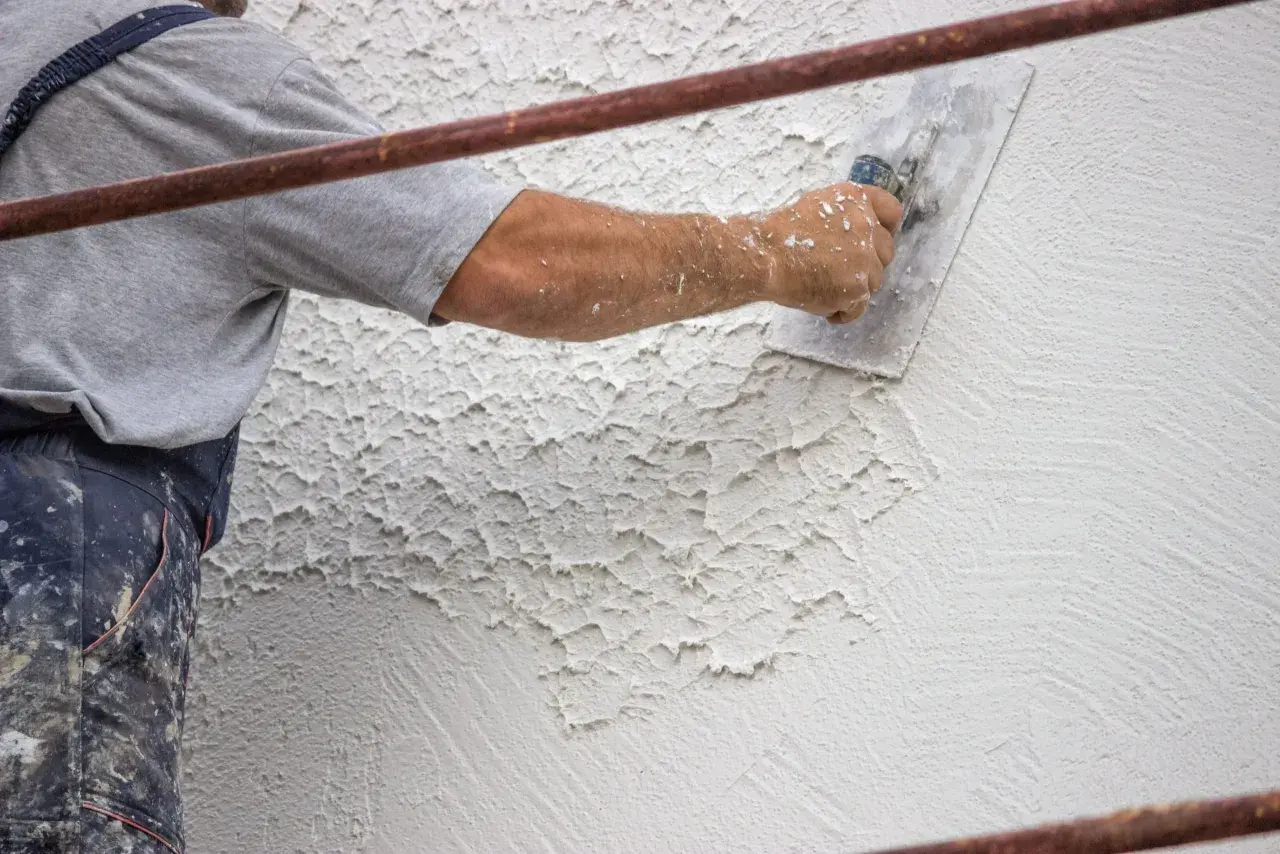 Mano di un operaio che applica intonaco rustico con una spatola, creando una texture grezza e irregolare su una parete.