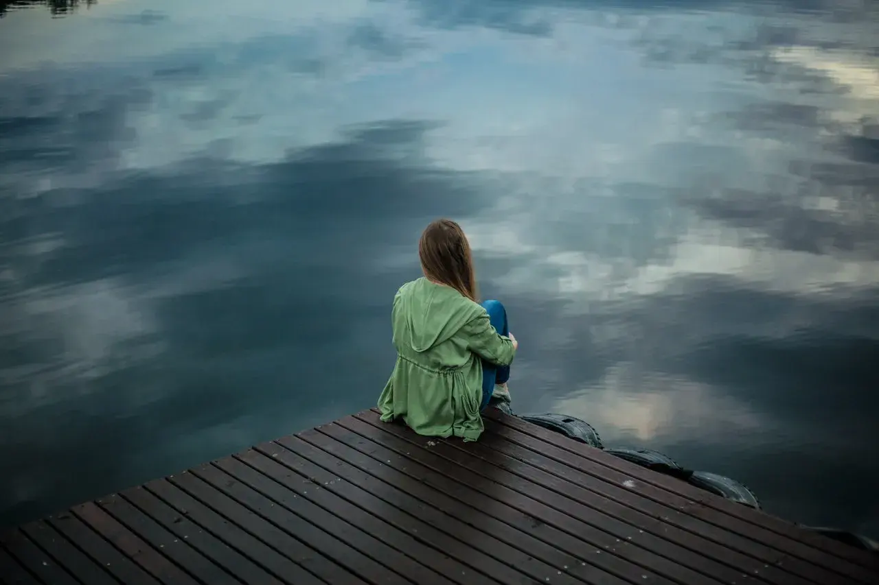 Ein Mädchen sitzt allein auf einem Holzsteg am Wasser. Sie wirkt nachdenklich, als ob sie nicht alleine sein kann.