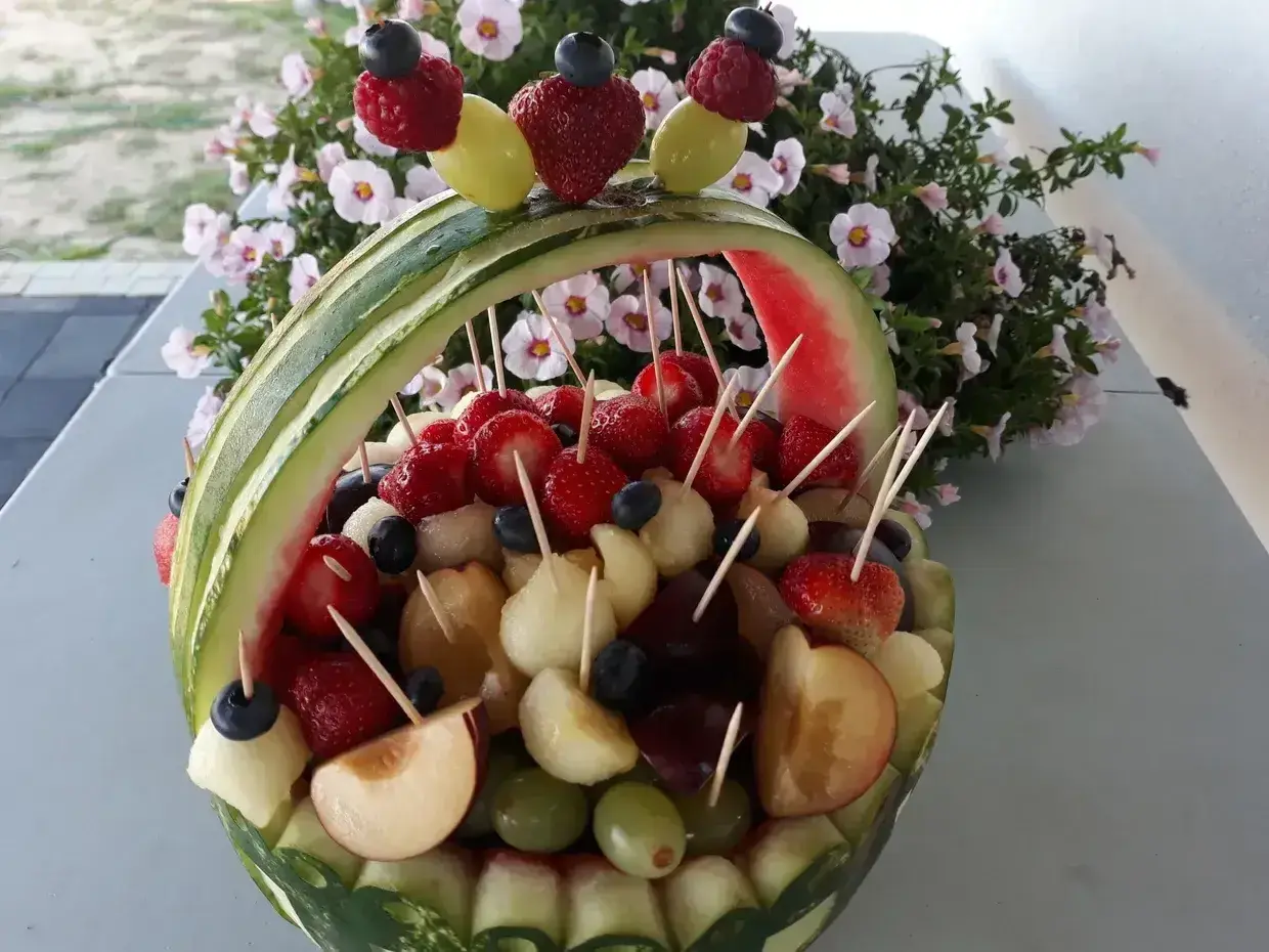 Koszyk z arbuza wypełniony owocami: truskawkami, winogronami, melonem i śliwkami. Idealne owoce na wesele, ozdobione malinami i borówkami.