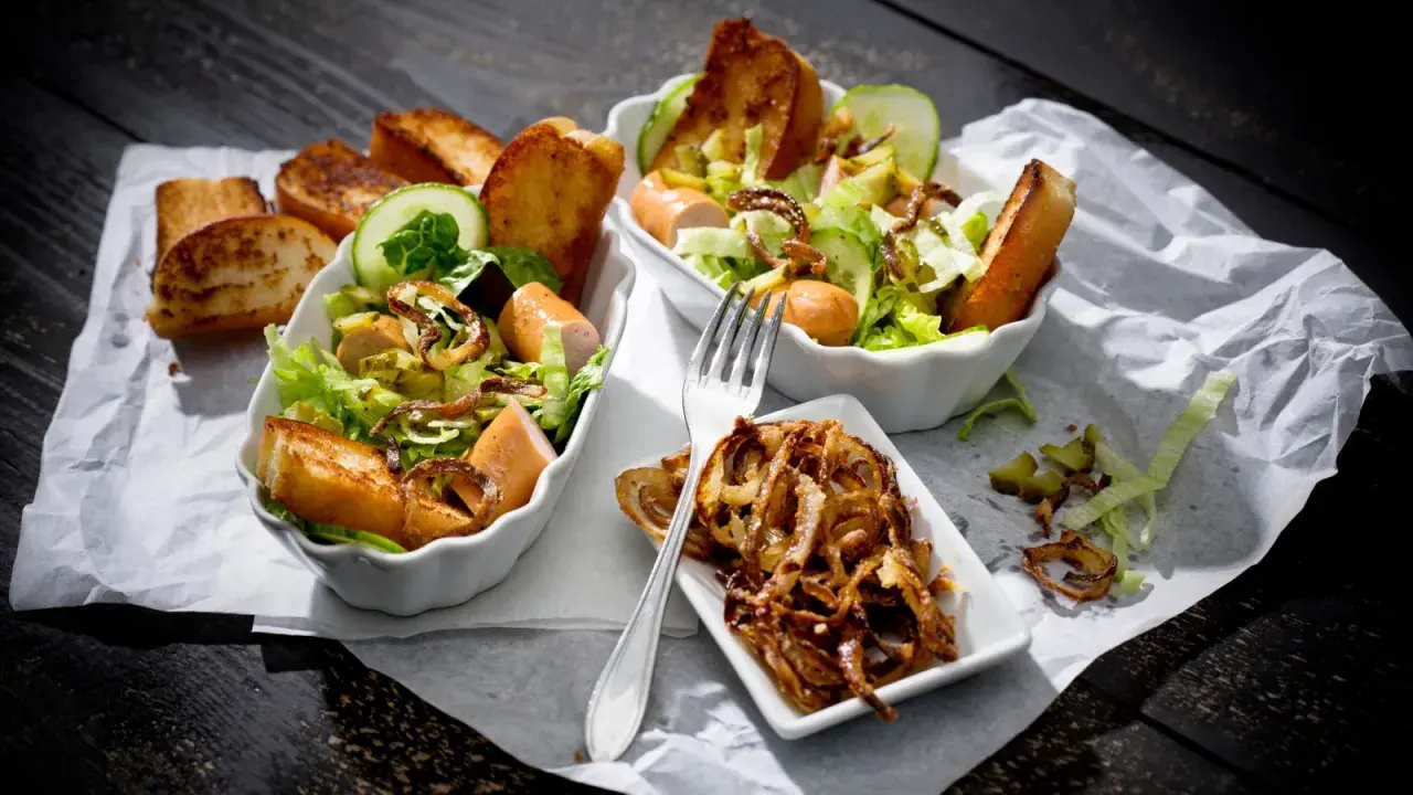 Zwei Schüsseln mit Hot Dog Salat, garniert mit Gurkenscheiben, geröstetem Brot und knusprigen Zwiebeln.
