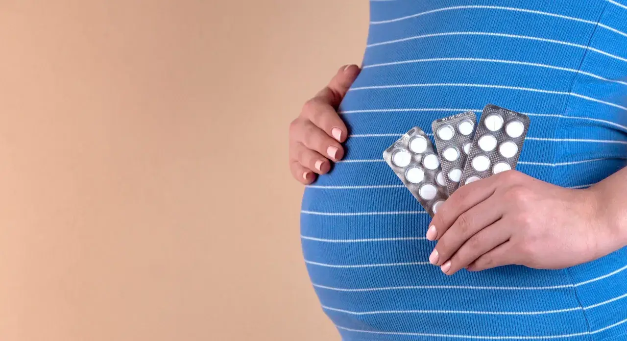 Gravid kvinna håller upp tre paket med piller, troligen gravid vitaminer, mot sin blårandiga klänning.