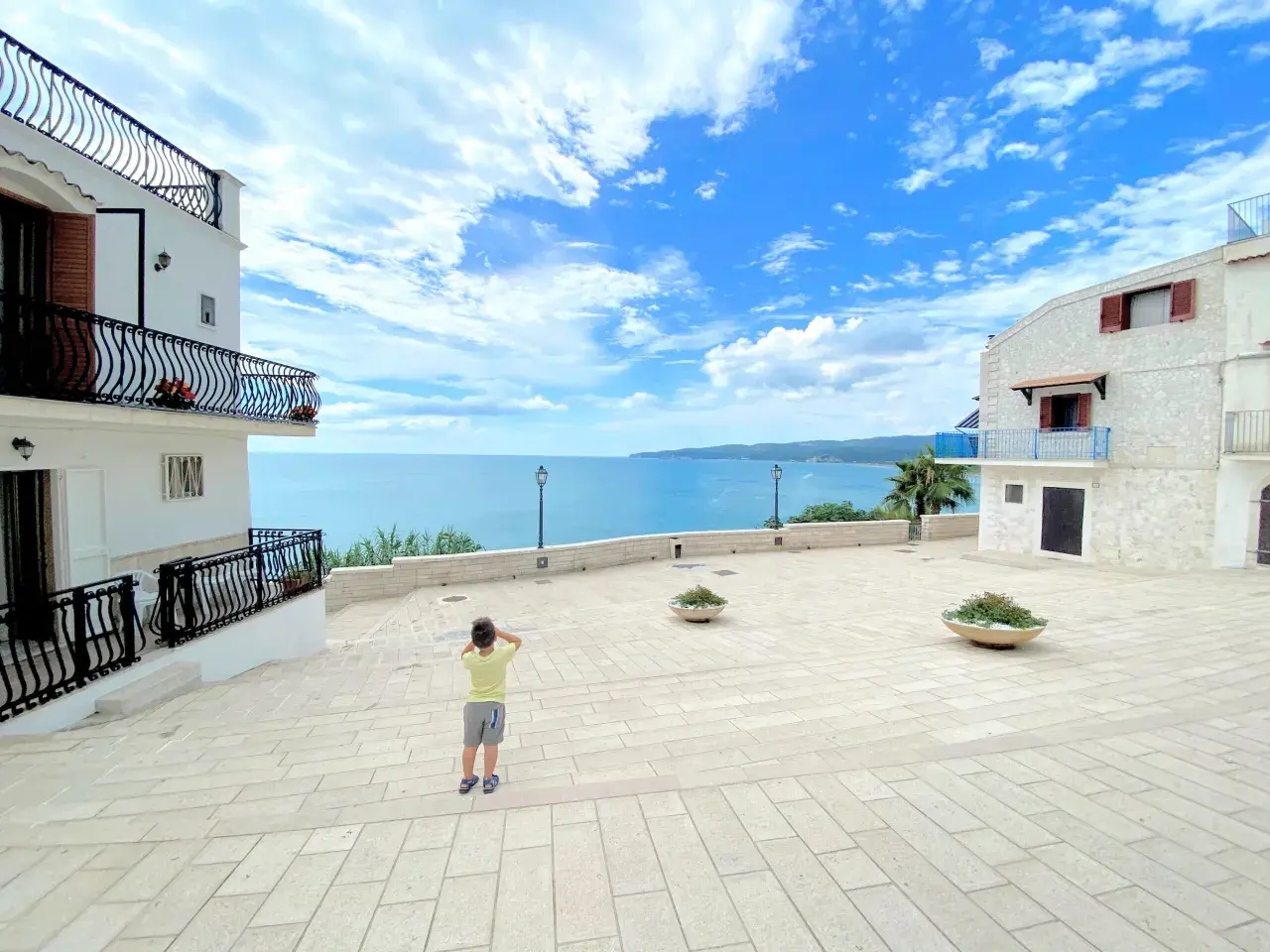 Bambino ammira il mare azzurro dalla piazzetta Petrone a Vieste, con case bianche e cielo nuvoloso.