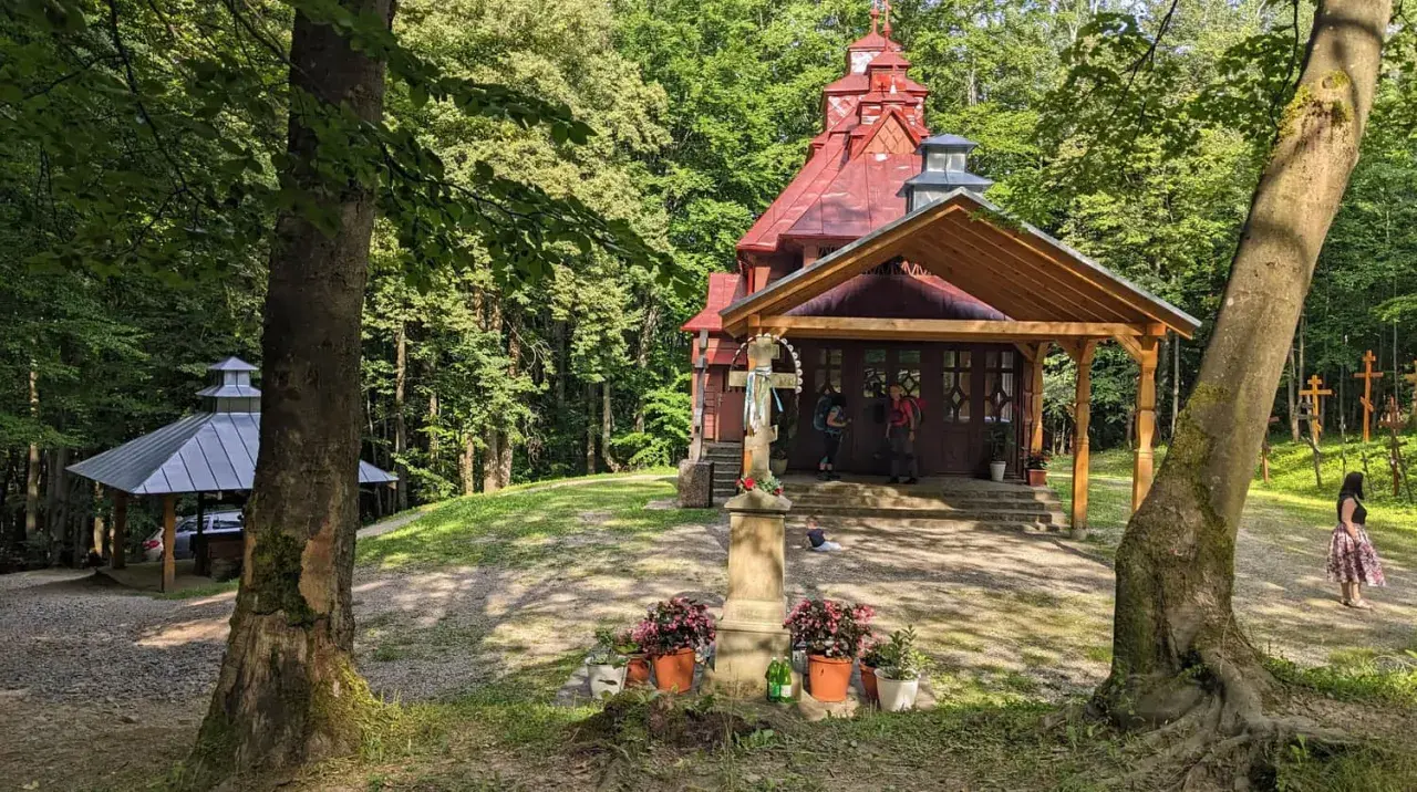 Kapliczka na górze Jawor otoczona lasem. Przed wejściem krzyż z kwiatami, obok wiata i cmentarz krzyży.