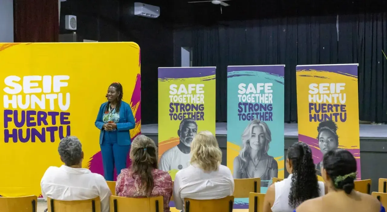 Een spreker staat voor een gele banner met "SEIF HUNTU FUERTE HUNTU". Achter haar staan banners met "SAFE TOGETHER STRONG TOGETHER". Dit is een evenement voor hss sw jeugd.