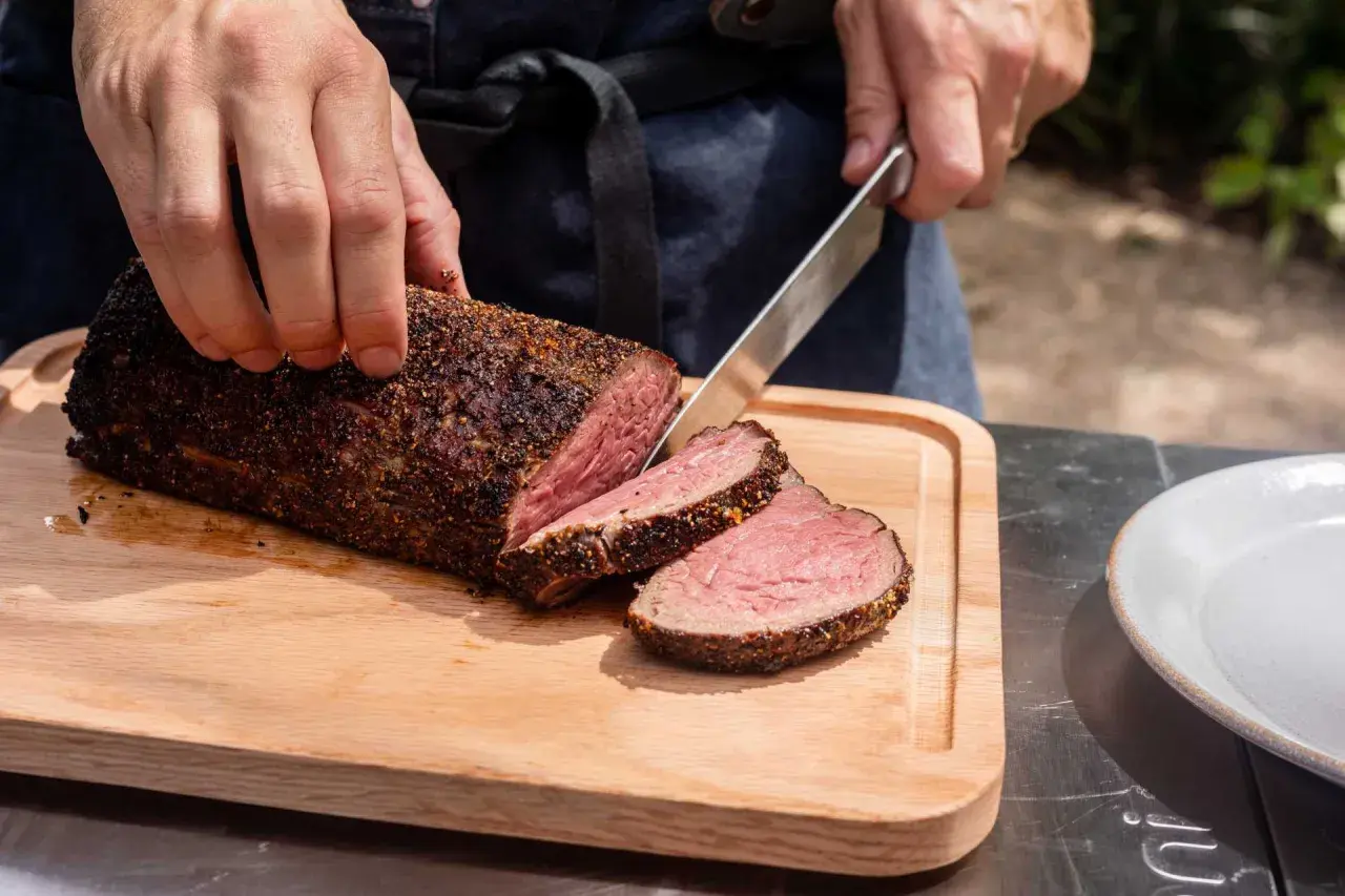 Un chef tranche un rôti de filet mignon parfaitement cuit sur une planche en bois.