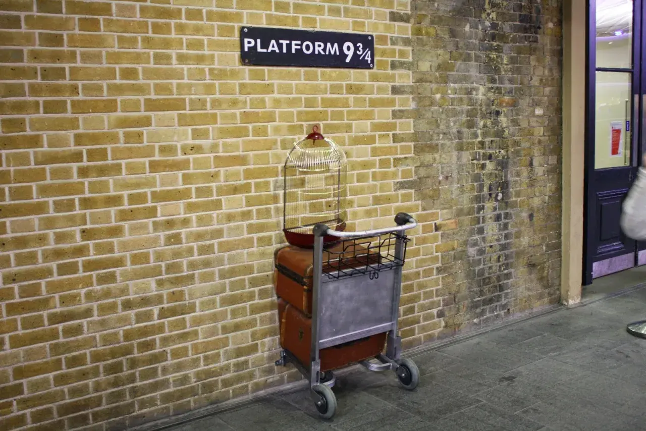 Wózek z bagażami i klatką dla ptaków przy ścianie z napisem "Platform 9 3/4" - magiczne miejsce jak z Kings Cross Harry Potter.