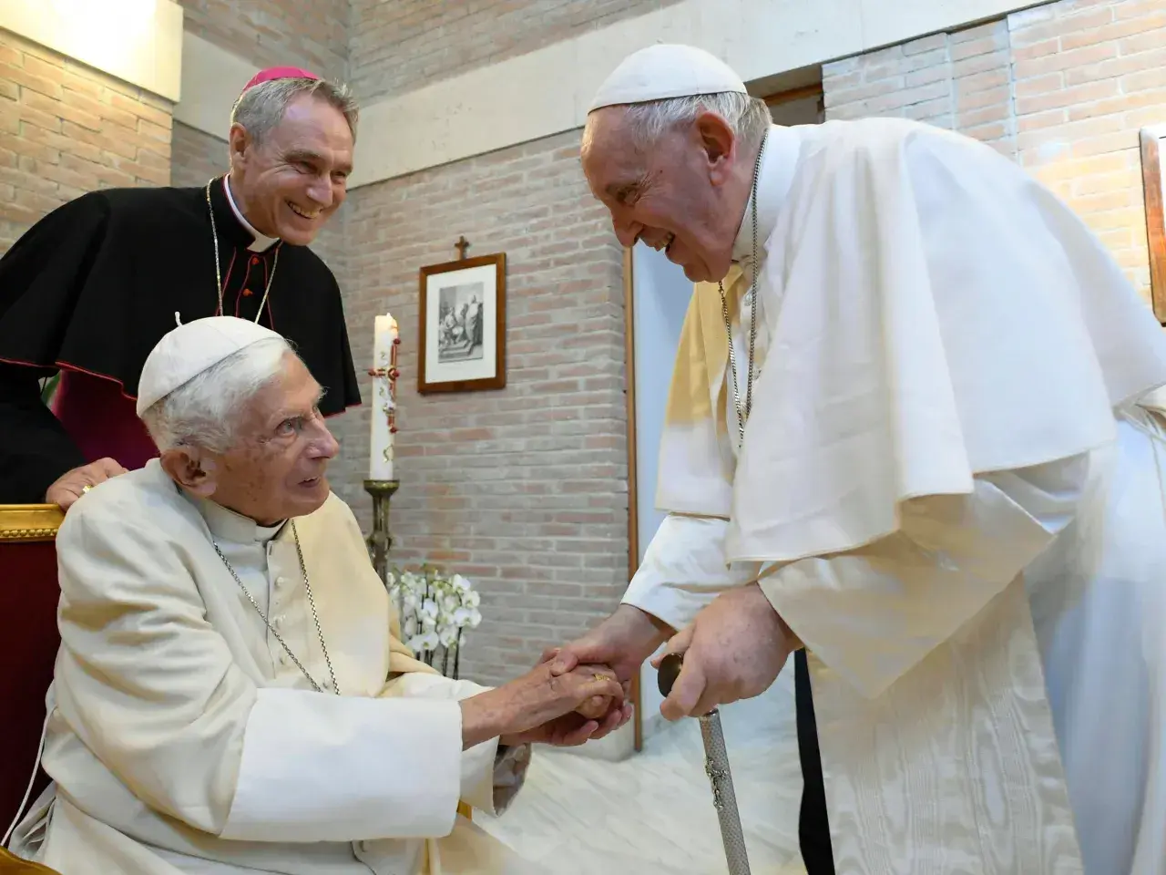 Papst Franziskus und ein Bischof besuchen Papst Benedikt. Sie reichen sich die Hände und lächeln.