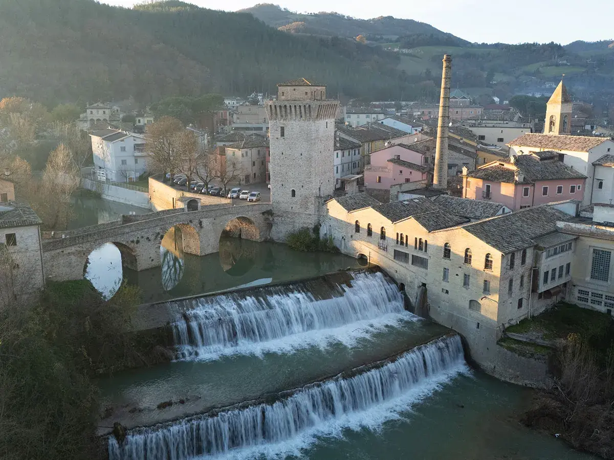 Cascata e ponte ad arco a Fermignano, vicino Urbino, con edifici storici e una ciminiera.