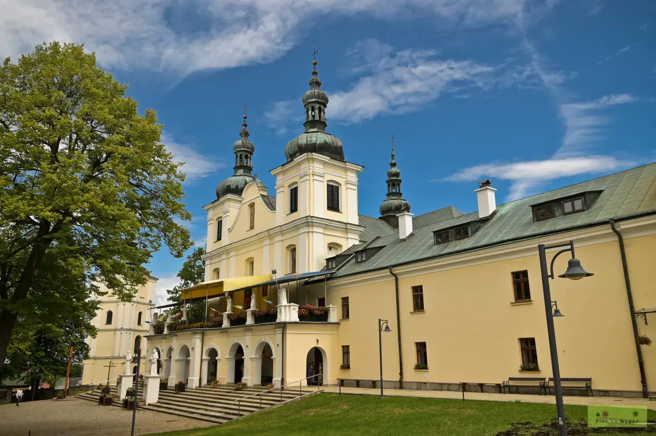 Jasny budynek sanktuarium z kopułami i krzyżami na wieżach, otoczony zielenią. Kalwaria Pacławska dróżki zapraszają do modlitwy.