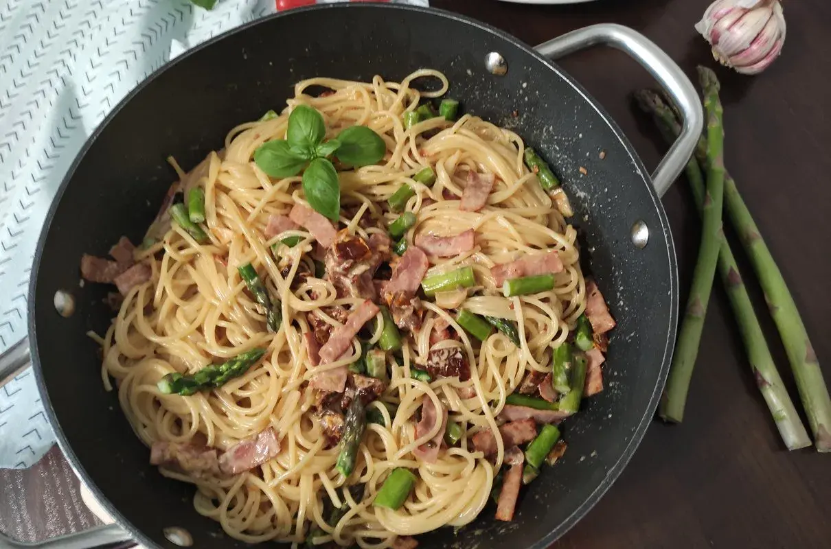 Pyszne spaghetti ze szparagami i boczkiem, udekorowane listkiem bazylii, czeka w patelni. Obok leżą świeże szparagi i czosnek.