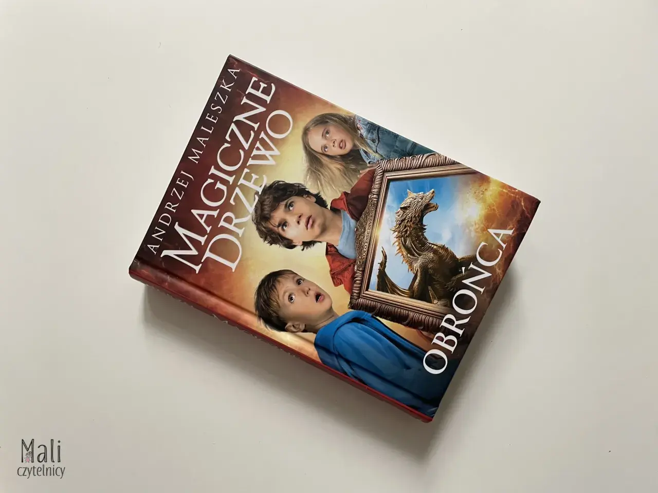 Okładka książki "Magiczne drzewo. Obrońca" z Andrzejem Maleszką. Trójka dzieci patrzy z zaciekawieniem na smoka w ramie.
