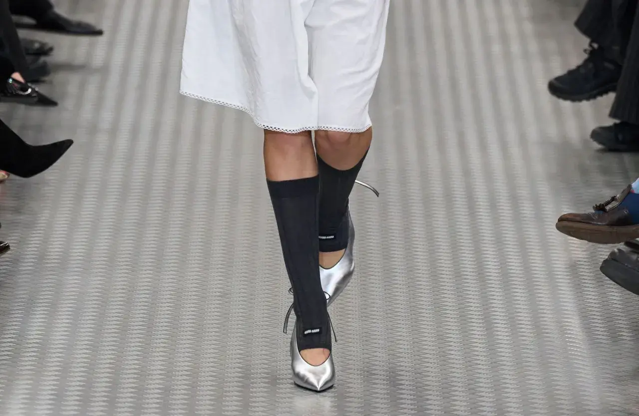 Une mannequin défile avec une tenue blanche et des chaussettes hautes grises, rehaussée par des chaussures argentées pointues.