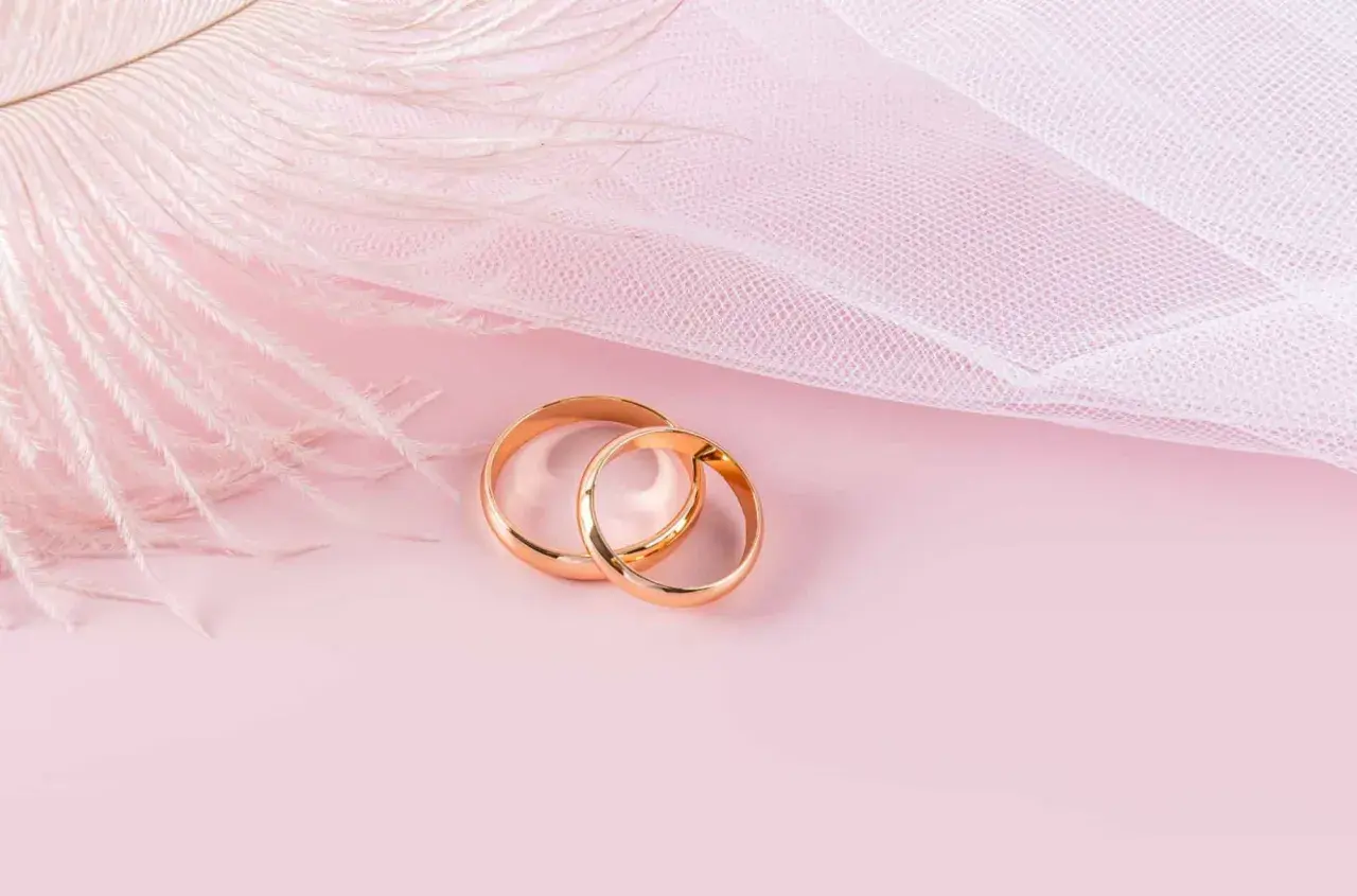 Dos anillos de oro rosa entrelazados, sobre un fondo rosa pálido con una pluma blanca y tela delicada.