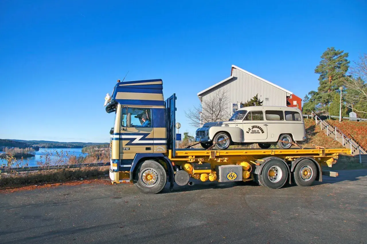En blågul lastbil transporterar en vit Volvo Raske herrgårdsvagn längs en sjö.