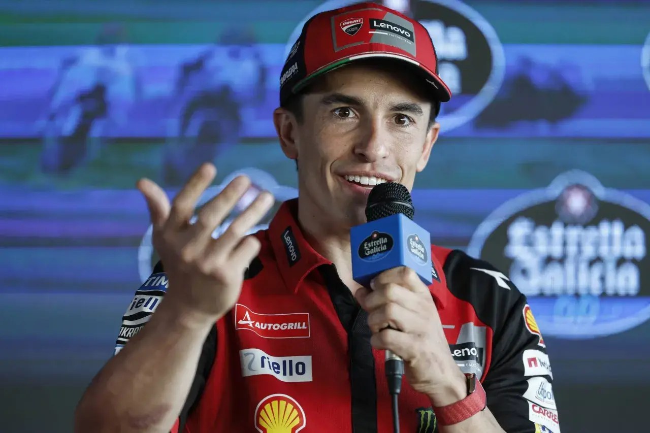 Marc Márquez, con su gorra roja y polo de Ducati, habla con un micrófono. Su edad se refleja en su energía y sonrisa.