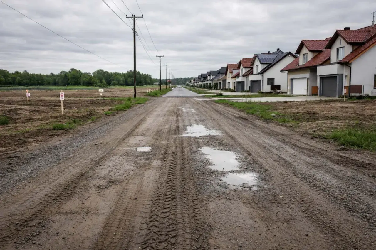 Błotnista droga polna prowadzi do szeregu nowych domów. Po lewej stronie widać niezabudowane działki z tabliczkami.