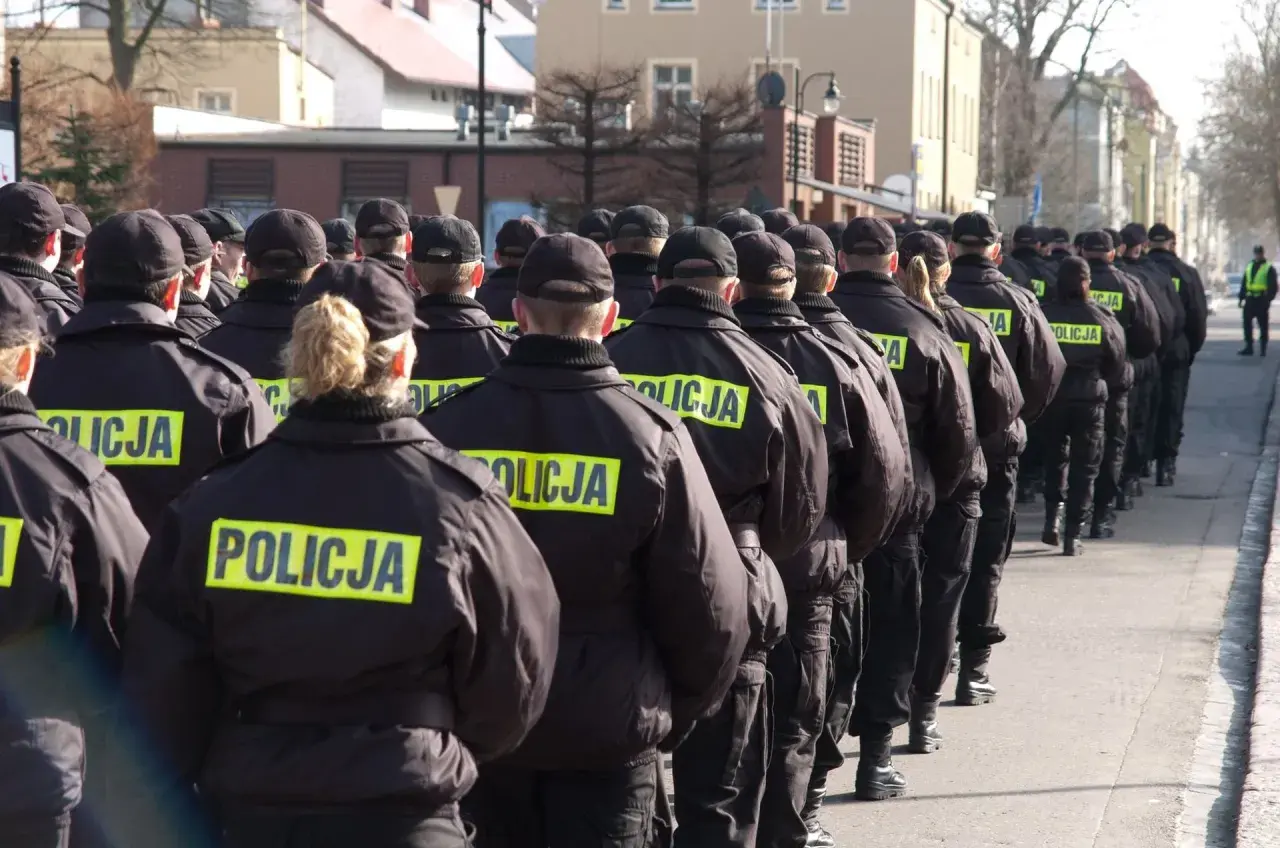 Policjanci w czarnych kurtkach z napisem "POLICJA" stoją w szeregu, gotowi do akcji. Widać, jak pracuje policja zmiany, przygotowując się do służby.