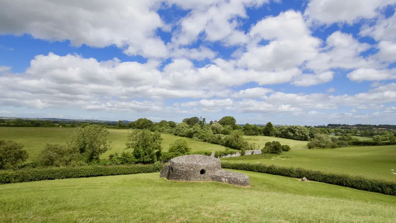 Antica struttura in pietra, forse un sito archeologico di Bru na Boinne, immersa in un paesaggio verdeggiante sotto un cielo azzurro con nuvole bianche.